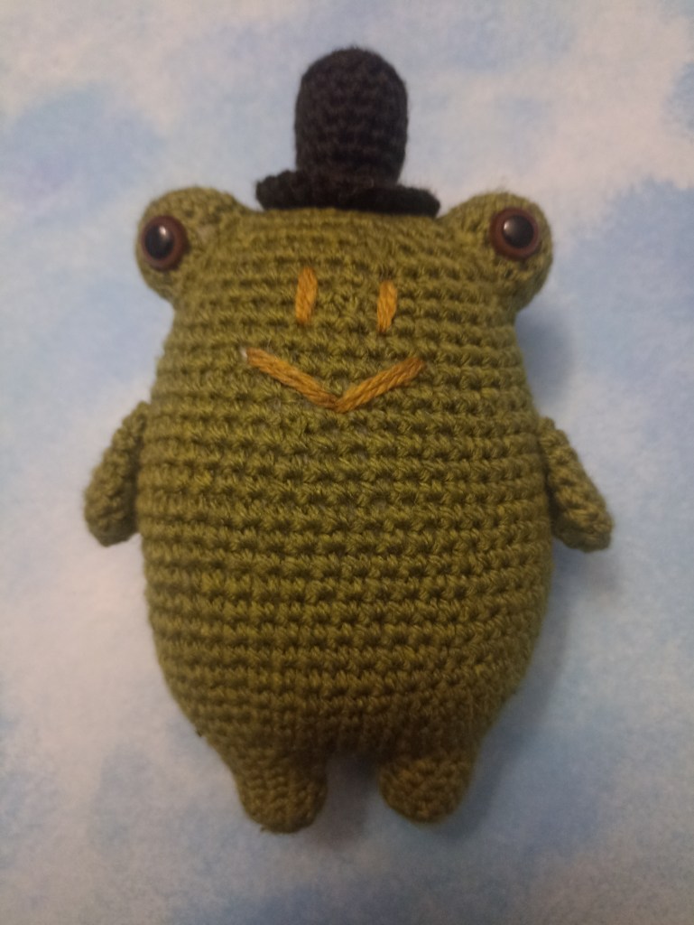A photo of Albie the Frog. A small crochet frog wearing a black top hat.